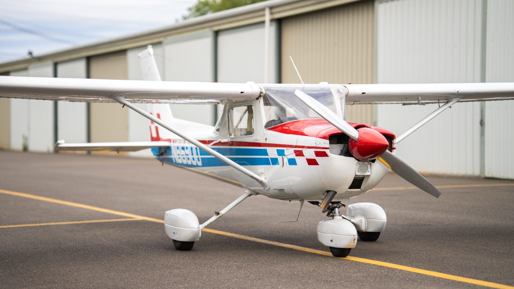Red, white and blue 1976 Cessna 150M parked on the ramp beside a hangar row